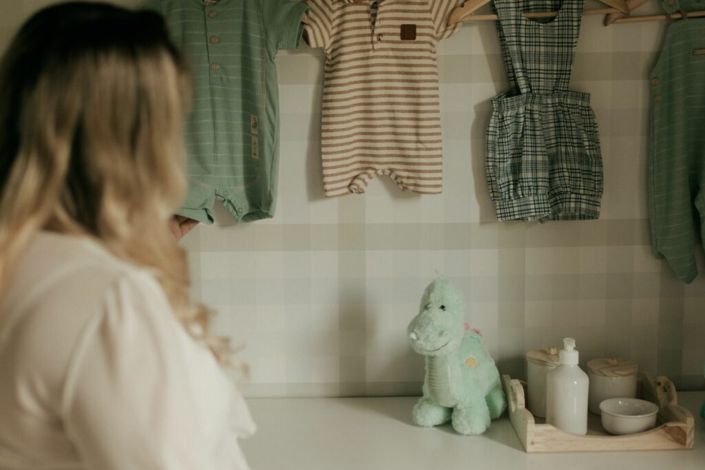 Future maman devant une commode blanche, vêtements de bébé suspendus au-dessus, chambre aux tons sauge