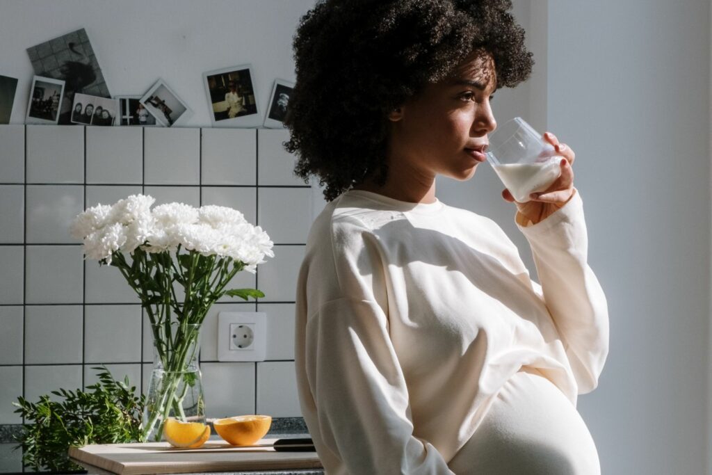 Femme enceinte dans une cuisine lumineuse, un verre à la main près d’un bouquet de fleurs blanches