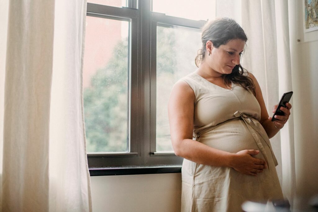 Femme enceinte près d’une fenêtre consultant son téléphone, une main sous le ventre