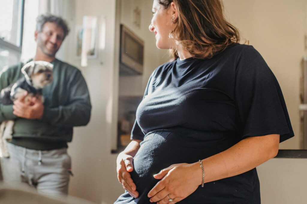 Femme enceinte souriante, une main sous le ventre, son compagnon en retrait avec leur chien