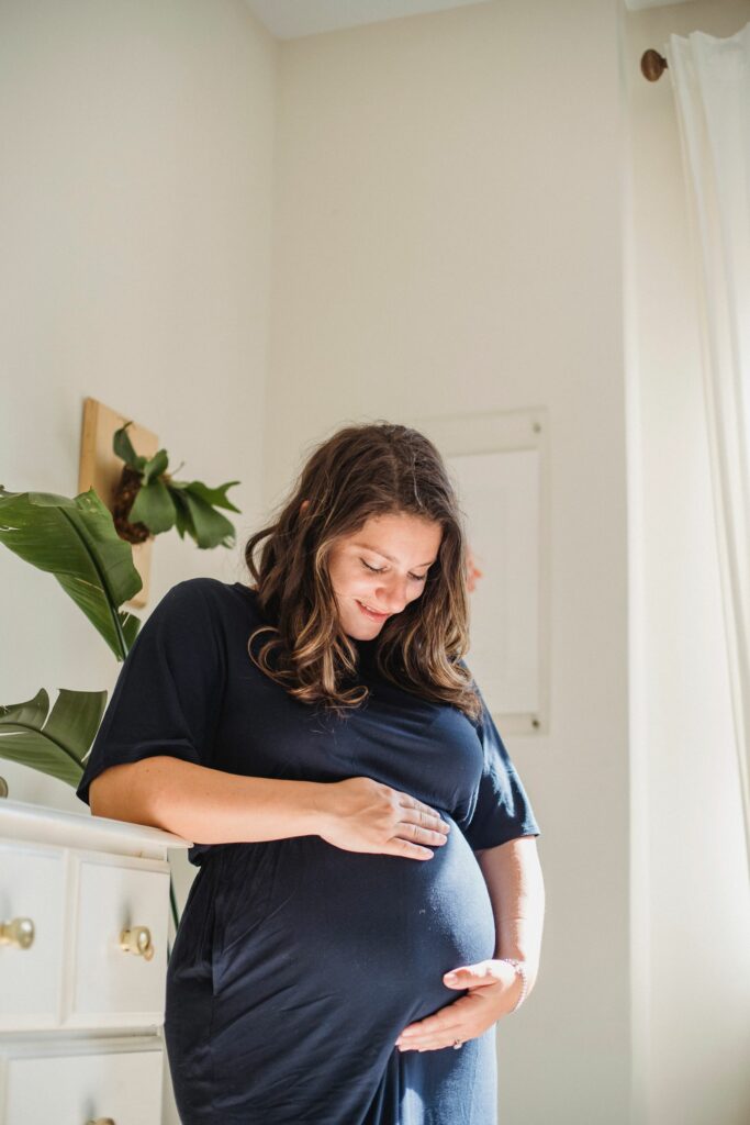 Future maman en robe bleue, mains autour de son ventre arrondi, dans une chambre lumineuse