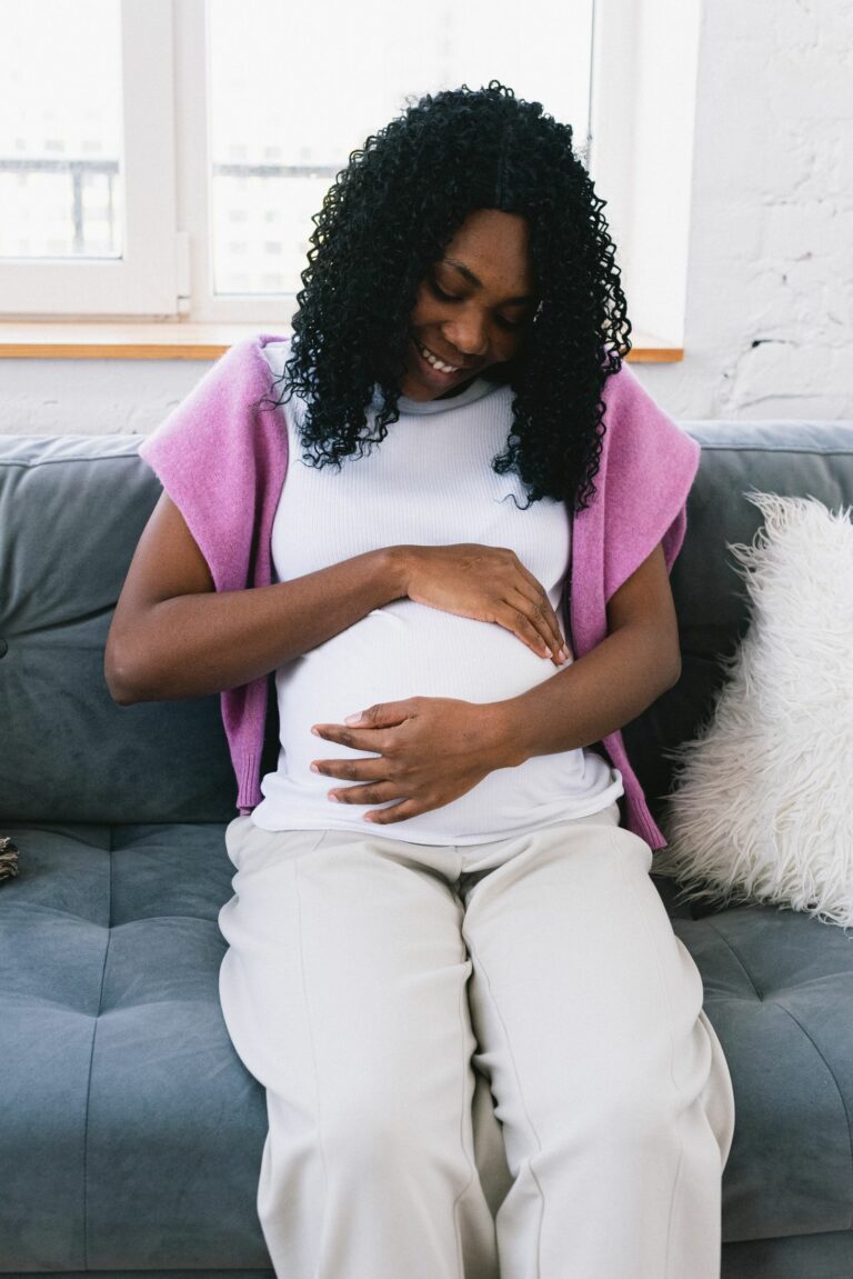 Future maman assise sur un canap&eacute;, mains pos&eacute;es sur son ventre, souriante