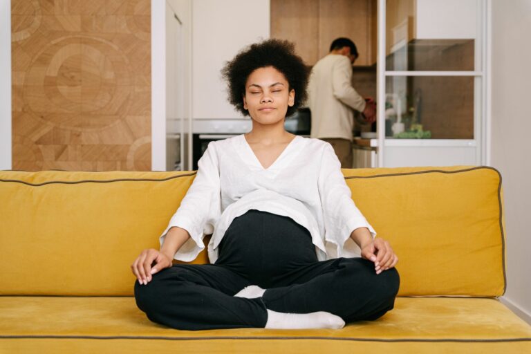 Future maman assise en tailleur sur un canap&eacute; jaune, les yeux ferm&eacute;s, sereine, son compagnon en arri&egrave;re-plan