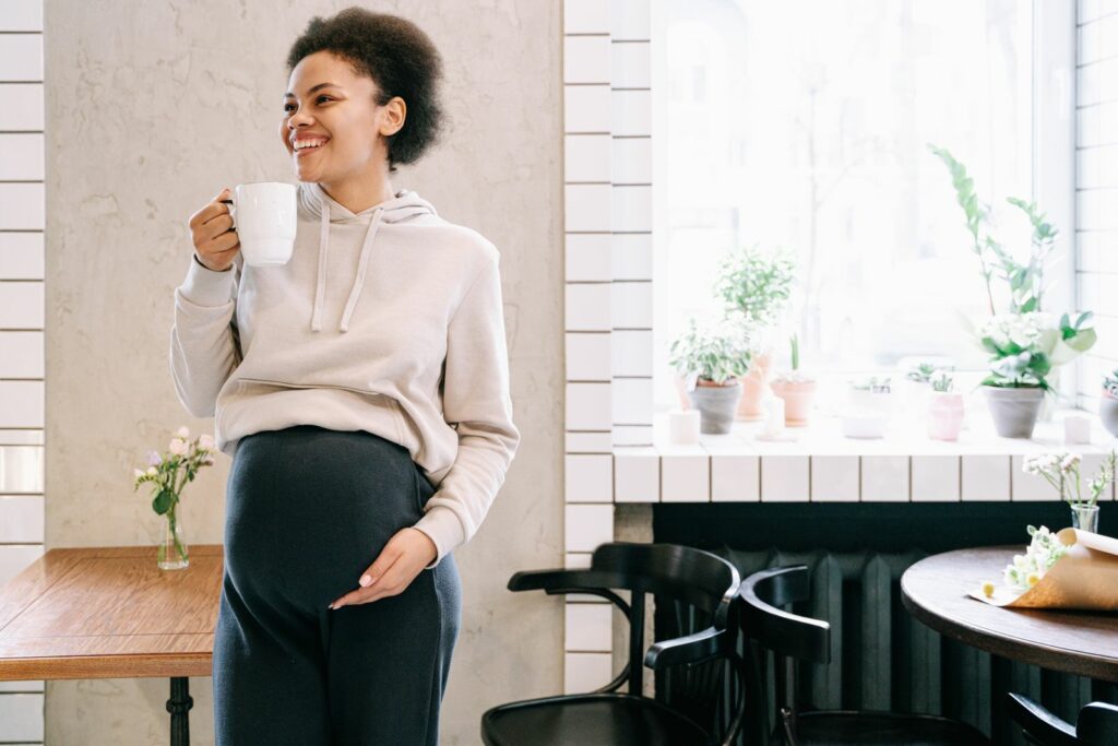Future maman souriante en sweat, une tasse à la main, détendue près d’une fenêtre lumineuse