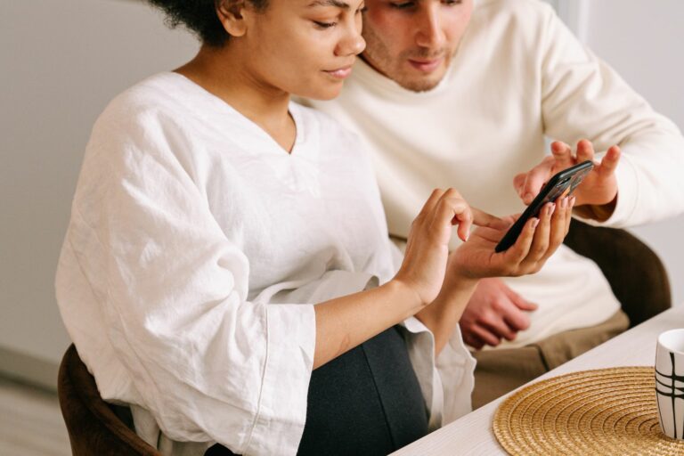 Un jeune couple choisit ensemble un prénom sur un téléphone, dans une cuisine lumineuse