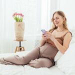 Future maman souriante installée sur son lit dans une chambre lumineuse, envoyant sa liste de naissance depuis son téléphone
