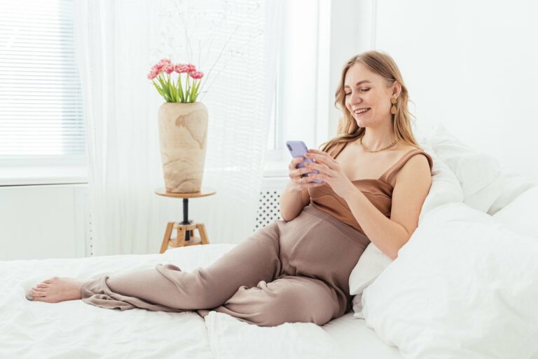 Future maman souriante installée sur son lit dans une chambre lumineuse, envoyant sa liste de naissance depuis son téléphone