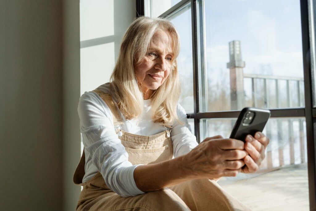 Grand-mère souriante assise près d'une fenêtre, consultant une liste de naissance sur son téléphone