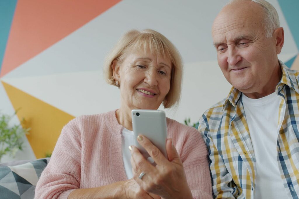 Couple de grands-parents souriants assis sur un canapé, regardant ensemble l'écran d'un smartphone