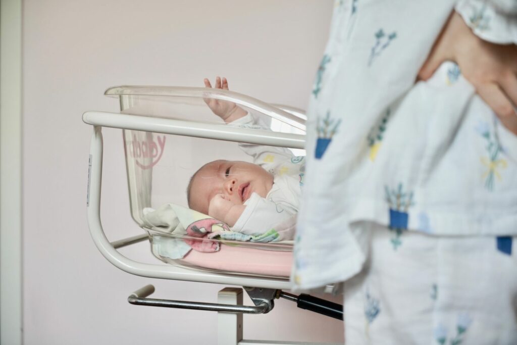 Nouveau-né éveillé dans un berceau transparent de maternité, sa maman debout à côté en chemise de nuit fleurie