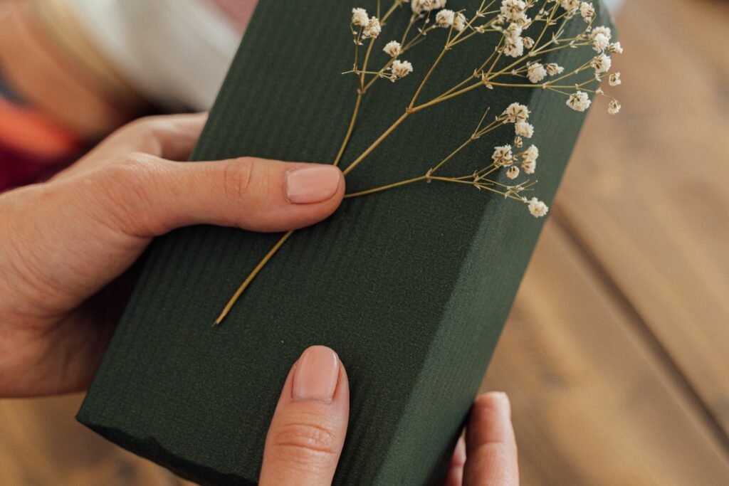 Mains tenant un cadeau de naissance emballé dans un papier vert, décoré d'un brin de gypsophile