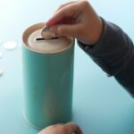 Une petite main glisse une pièce dans une tirelire turquoise posée sur une table