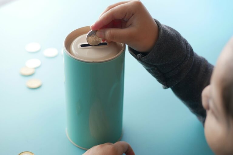 Une petite main glisse une pièce dans une tirelire turquoise posée sur une table