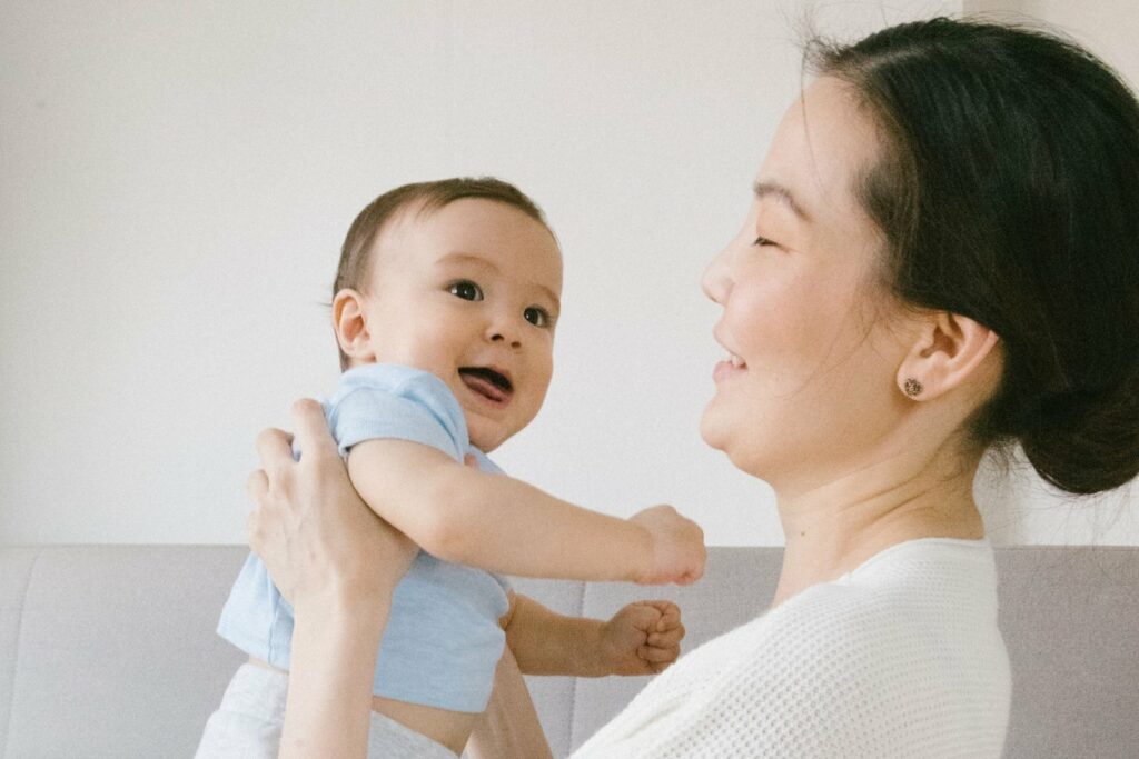 Une maman souriante soulève son bébé en body bleu ciel dans un salon clair