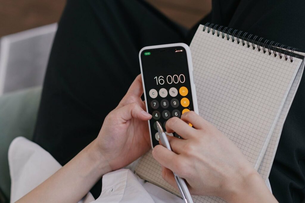 Des mains font un calcul sur la calculatrice d'un téléphone, un carnet à spirale posé à côté