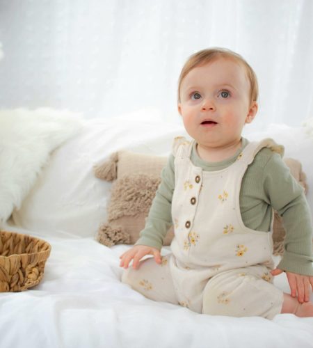 Charming baby in cozy outfit sits on a white bed surrounded by soft decor.